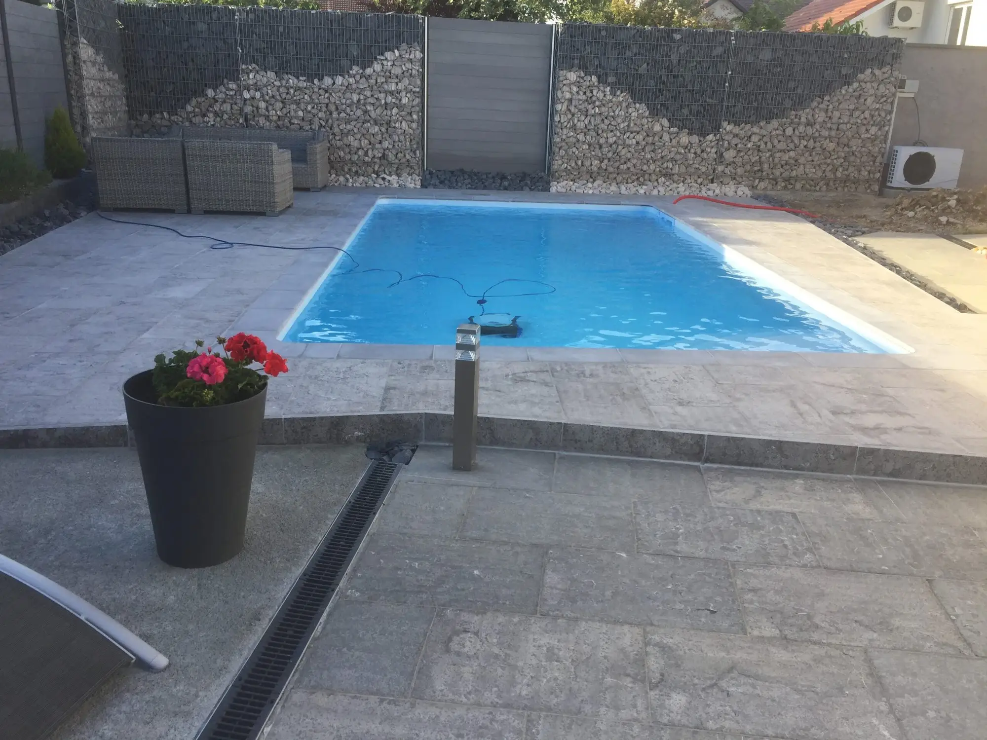 Plage de piscine en dallage clair avec pavés, mur en gabions et jardinière fleurie